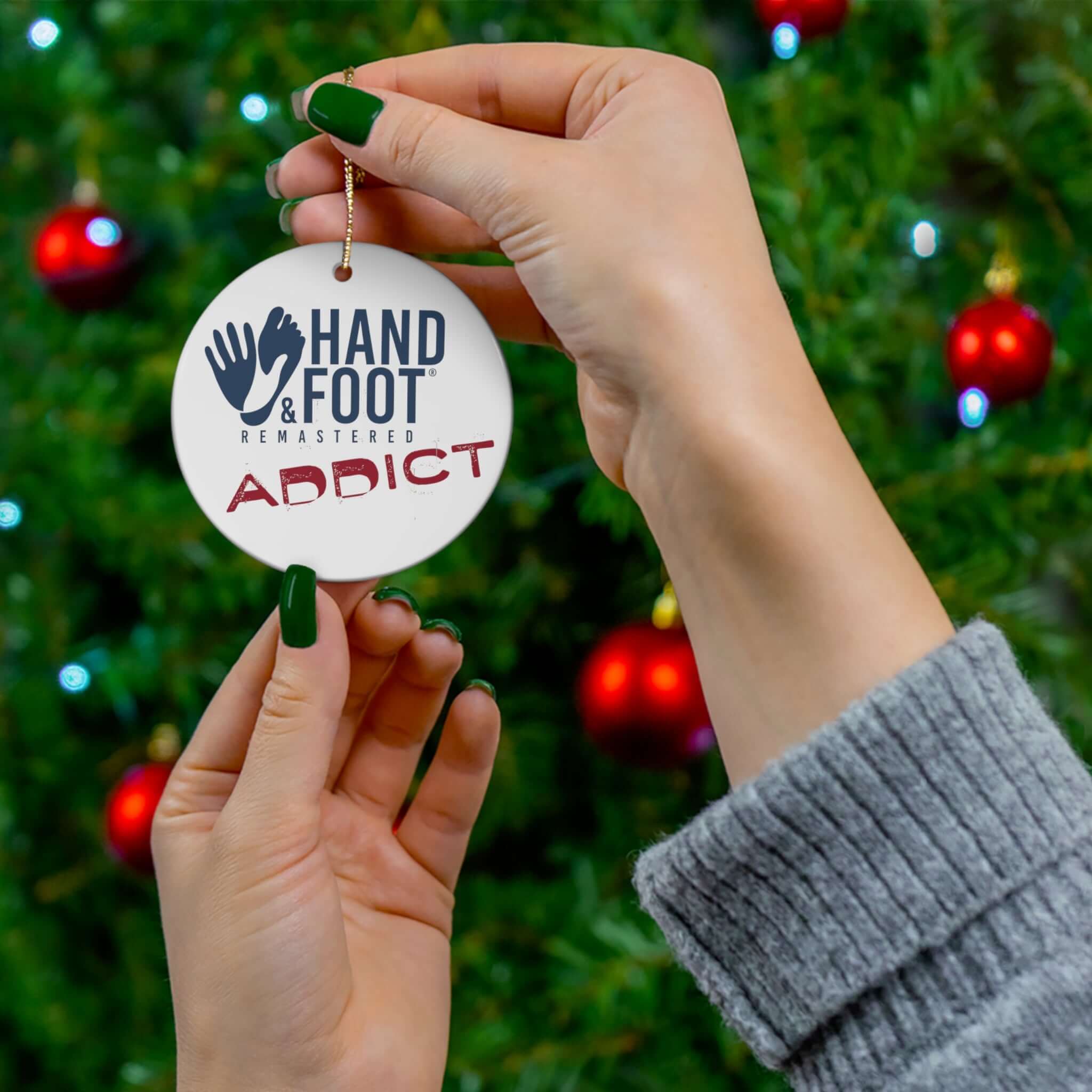 Person hanging Hand & Foot Addict ceramic ornament on a Christmas tree, showcasing festive decoration with red baubles and green branches.