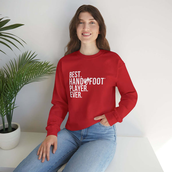 Unisex red sweatshirt with 'Best Hand/Foot Player Ever' text, worn by smiling person sitting indoors next to plant