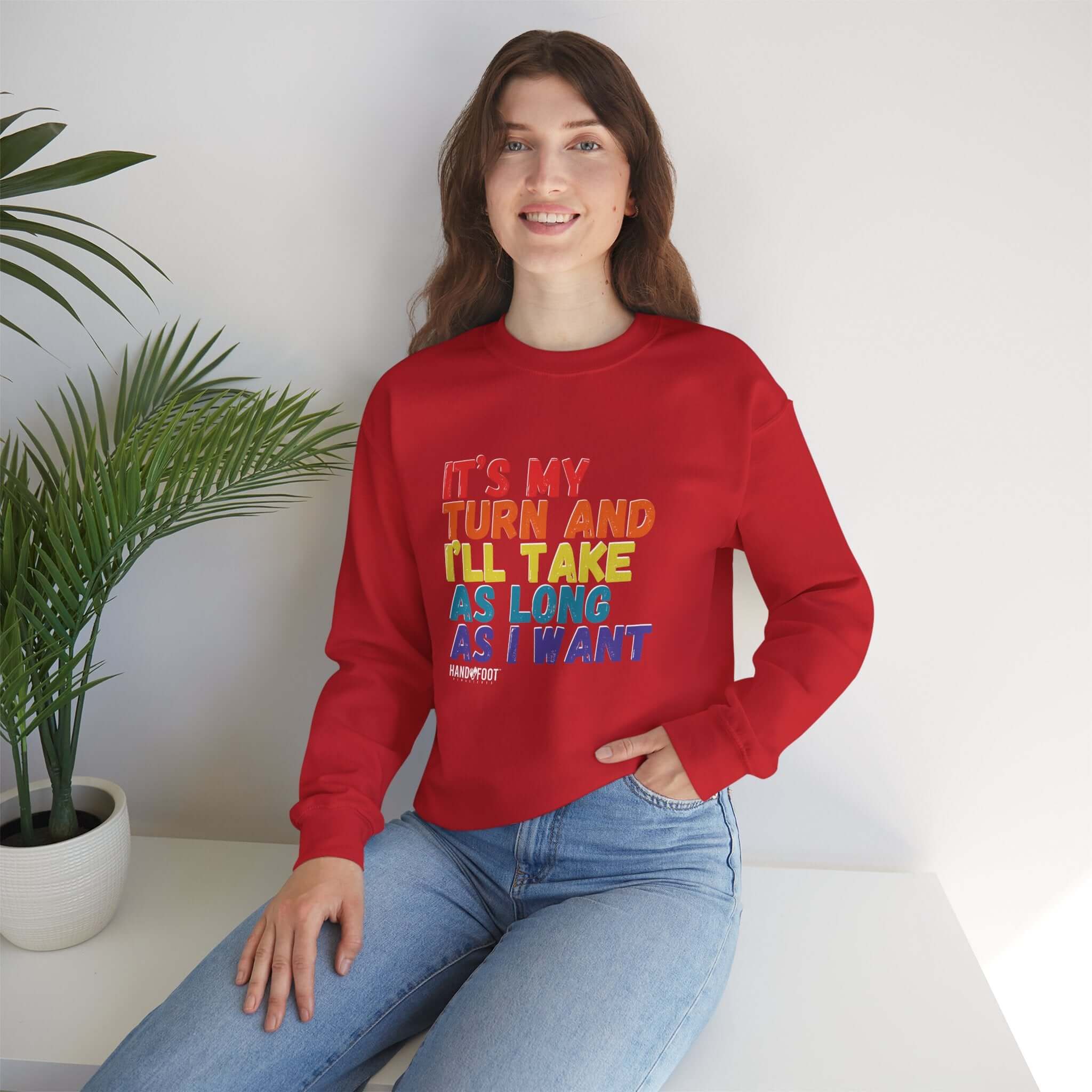 Woman wearing a red 'It's My Turn' unisex crewneck sweatshirt, sitting next to a plant, showcasing casual fashion style.