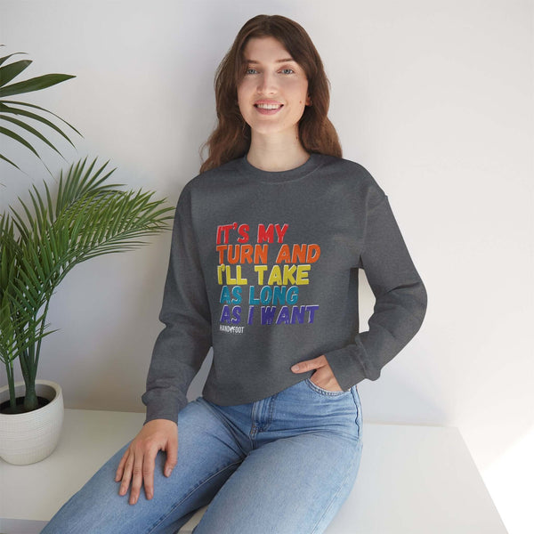 Model wearing 'It's My Turn' unisex sweatshirt, dark gray with colorful text, seated indoors with plant for a casual look.
