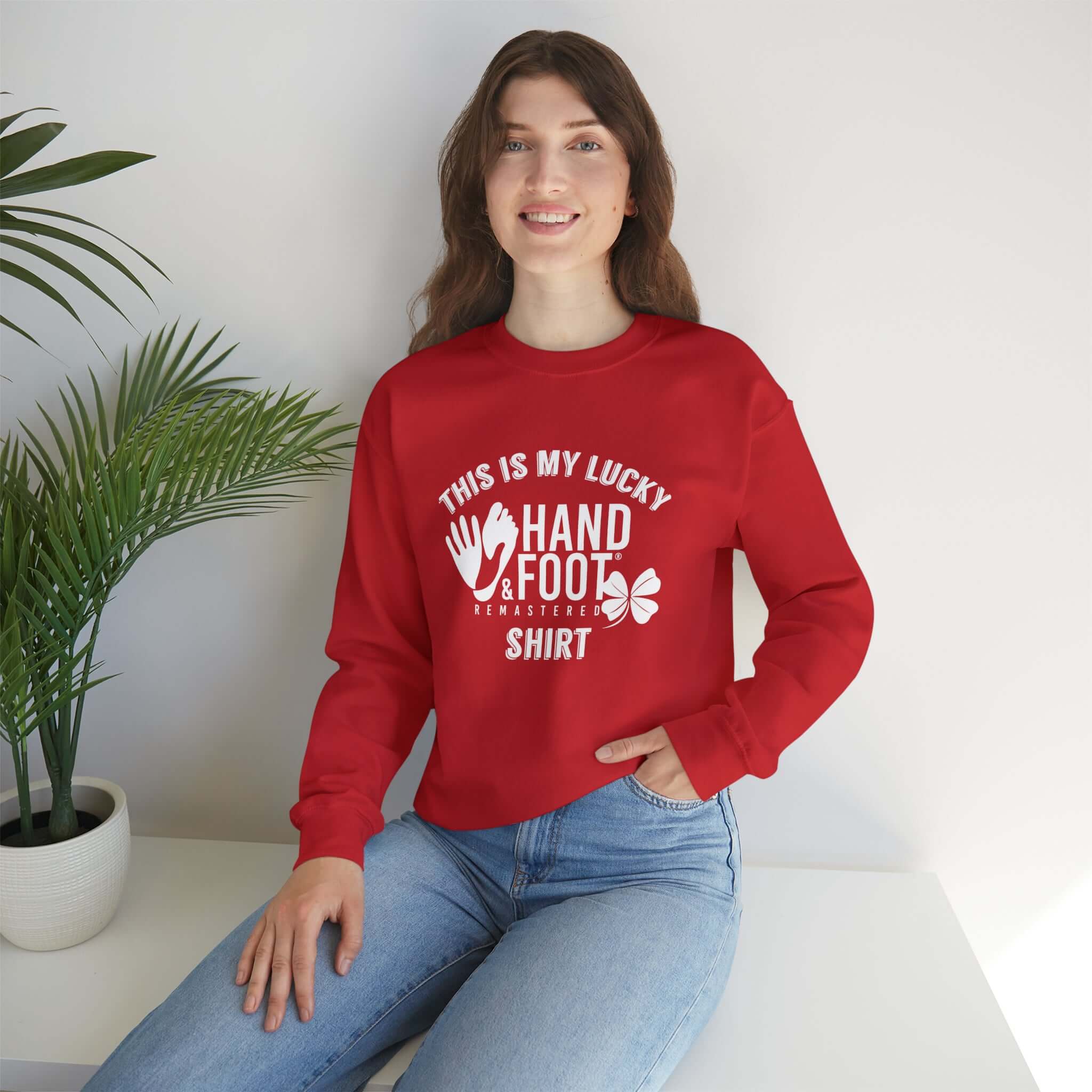 Smiling person wearing a red Lucky Unisex Heavy Blend Crewneck Sweatshirt with white graphic print, sitting near a plant.