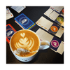Top view of latte art in a mug next to colorful card game sets on a table.