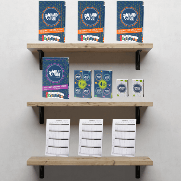 Hand & Foot Remastered Retail Starter Pack displayed on wooden shelves, featuring game editions, score pads, and action packs.