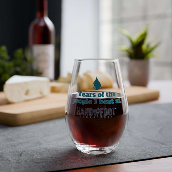 Stemless wine glass with 'Tears of the People I Beat at Hand & Foot' slogan, holding red wine, on a table set for casual dining.